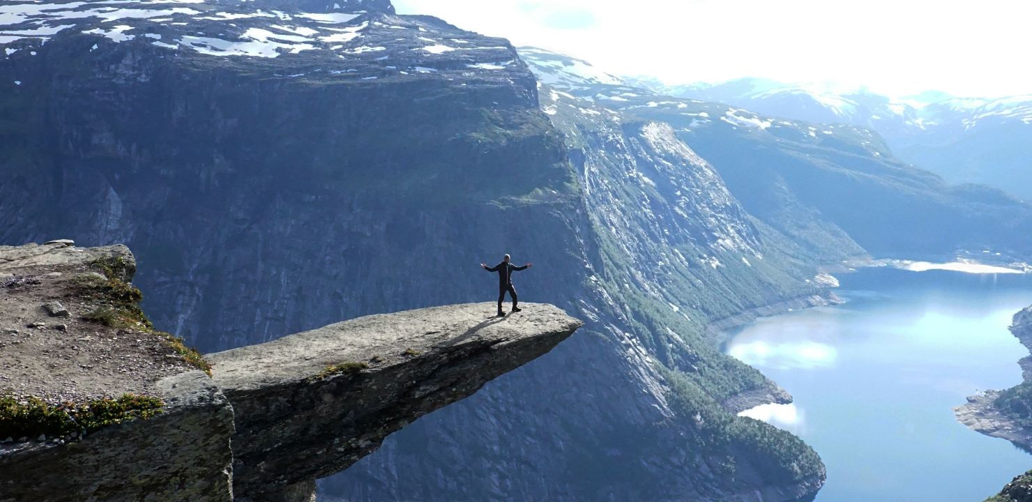 Manny standing with arms outstretched on a rock ledge above a deep blue fjord
