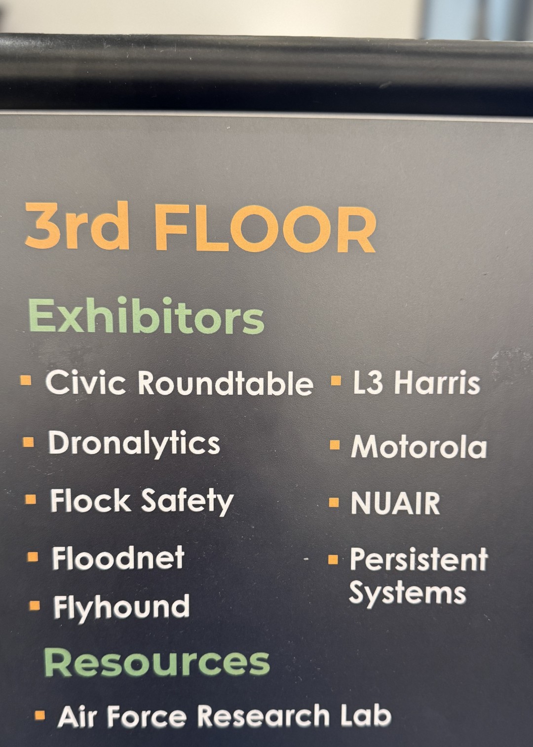A sign listing exhibitors at the conference with L3Harris, Motorola, Flock Safety mentioned along with Flyhound